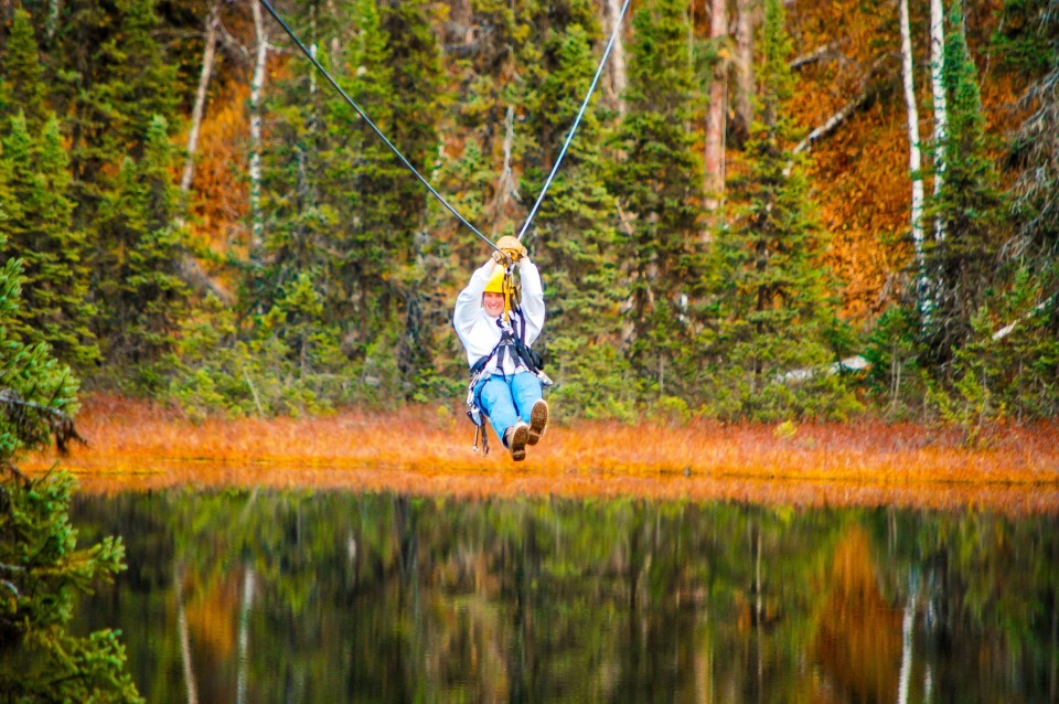 A woman rushes through the air over a lake on a zipline in an Alaska autumn forest setting.