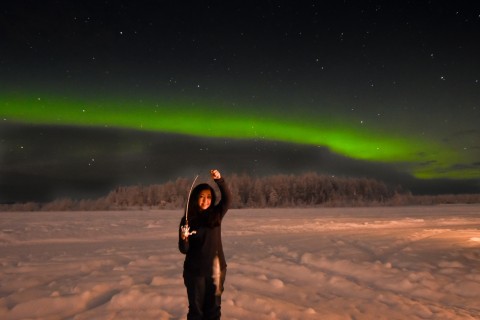 Vibrant green Northern Lights shine above someone who holds up a fish they caught while ice fishing.
