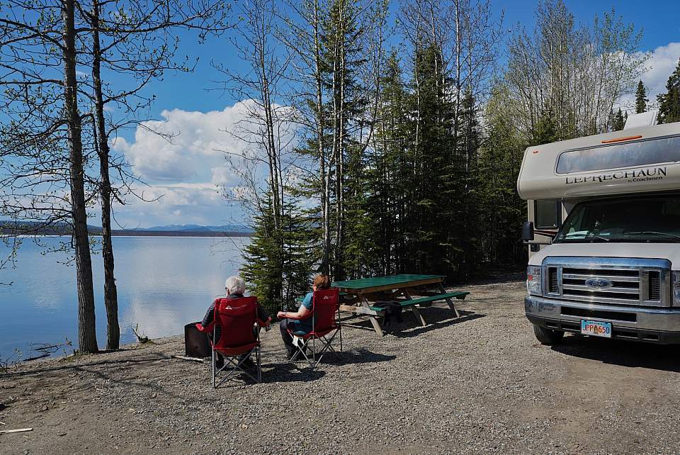 A couple sits on camp chairs in front of their RV overlooking at lake