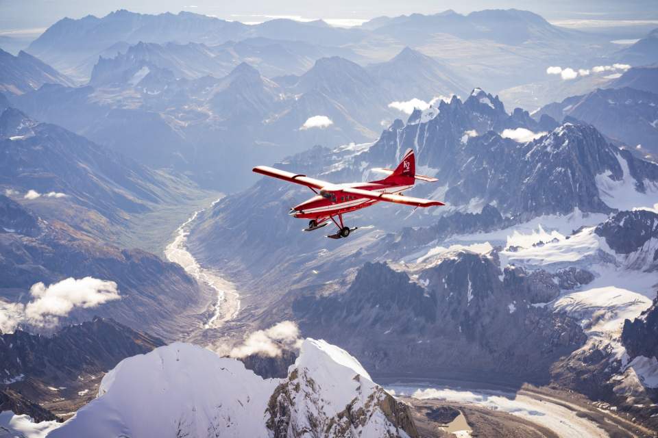 A small, red plane soars above a mountainous Alaskan landscape