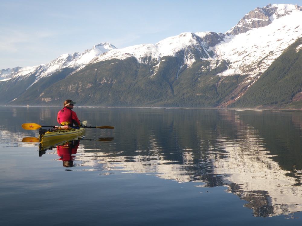 A person in a kayak on the water with snow capped mountains in the background