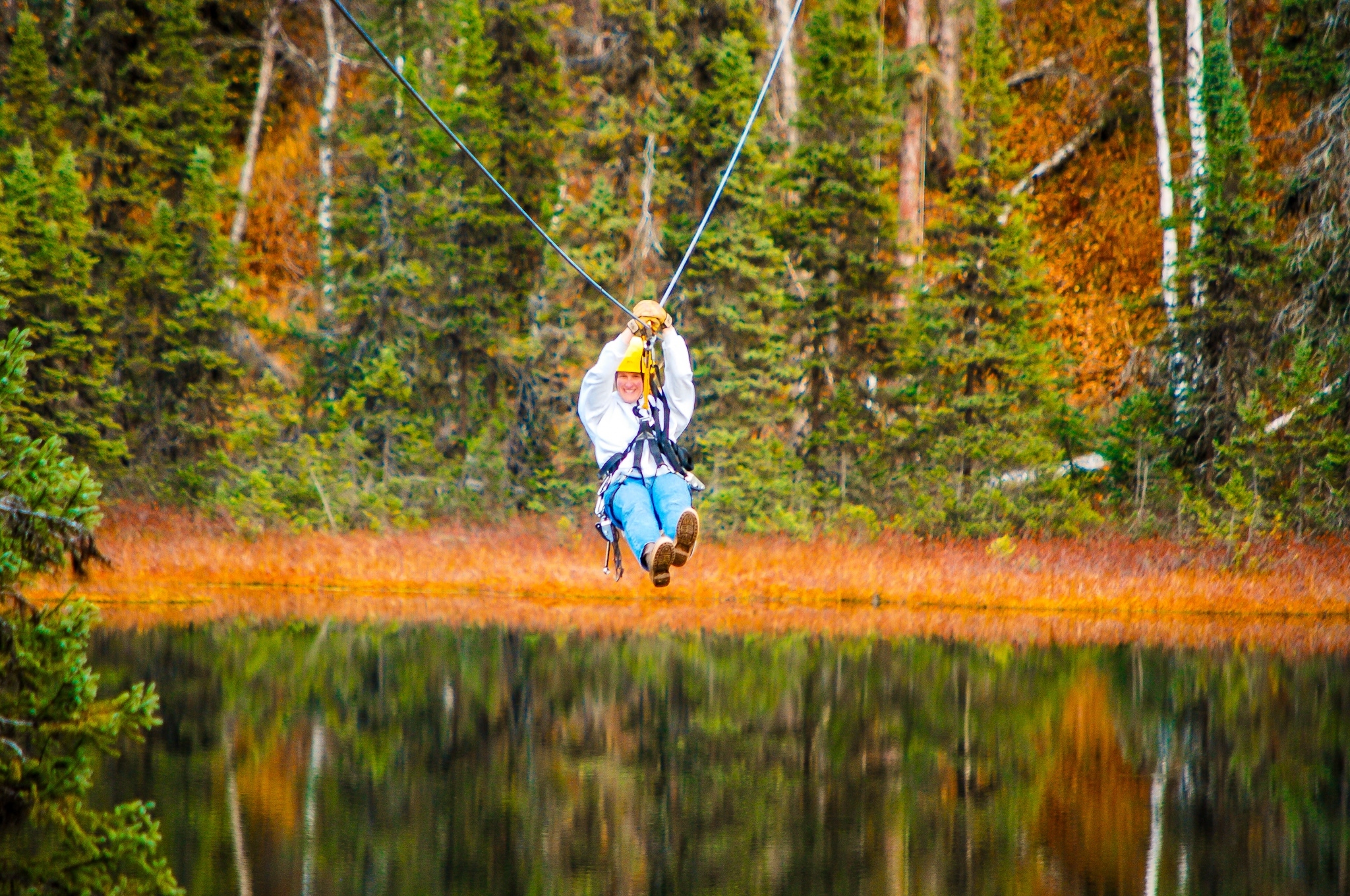 A woman rushes through the air over a lake on a zipline in an Alaska autumn forest setting