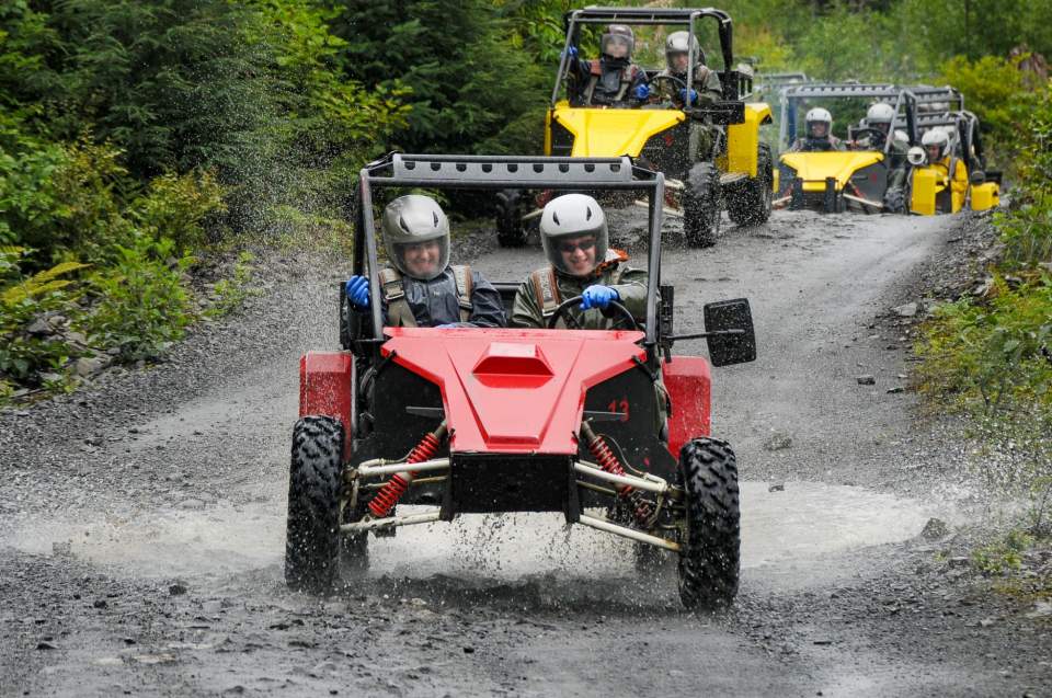 People in Tomcars race down a muddy forest trail.