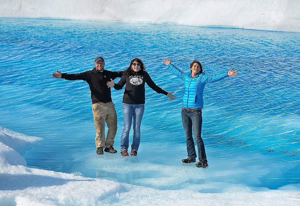 Three people pose with arms outstretched while they stand on the vibrant blue Knik Glacier.