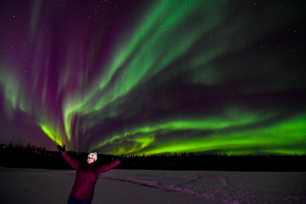 Woman stands under green bands of aurora borealis at Aurora Pointe near Fairbanks