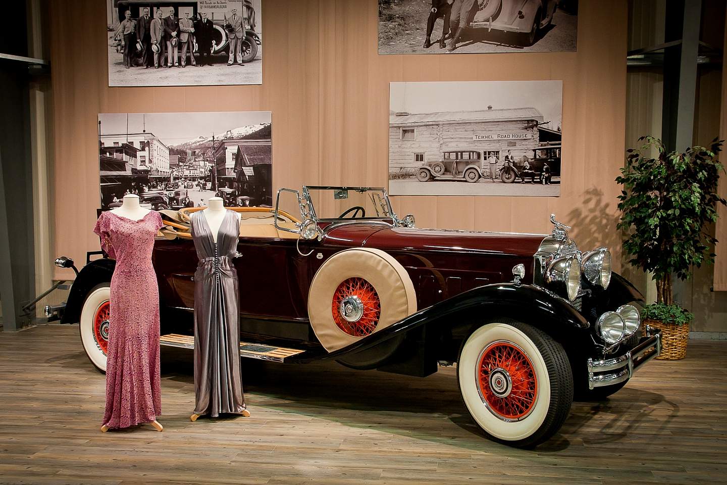 Two vintages dresses stand on display in front of a black and red vintage car with old photos displayed on the wall behind it