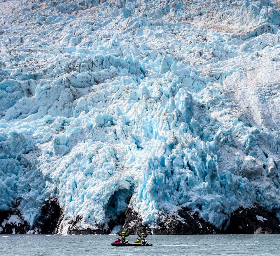 Two jet skiers in dry suits and helmets pose on the water in front of a glacier.