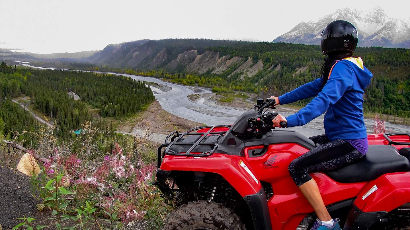 A woman on a red ATV overlooks an Alaskan valley with a river running through it.