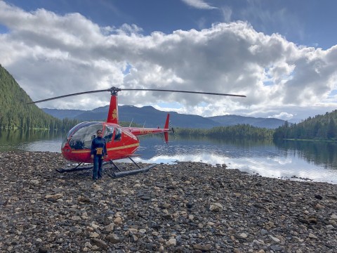 Pilot stands by their red helicopter that is landed on a rocky beach with mountains in the background.