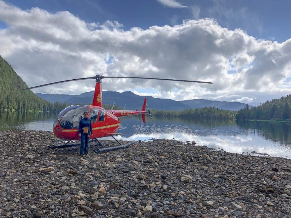 Pilot stands by their red helicopter that is landed on a rocky beach with mountains in the background.