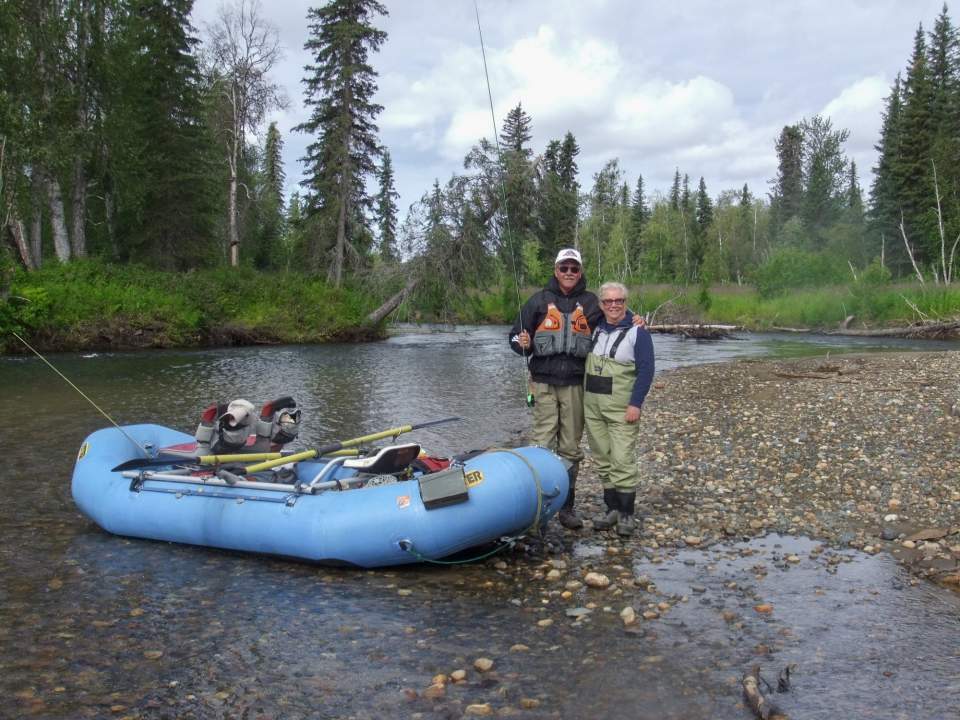 A couple in waders holding fishing poles poses by their raft next to the water.