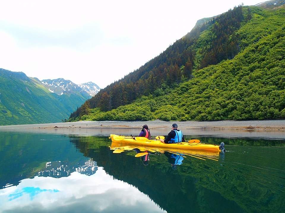 Two people in a yellow kayak glide on the water surrounded by lush mountains.