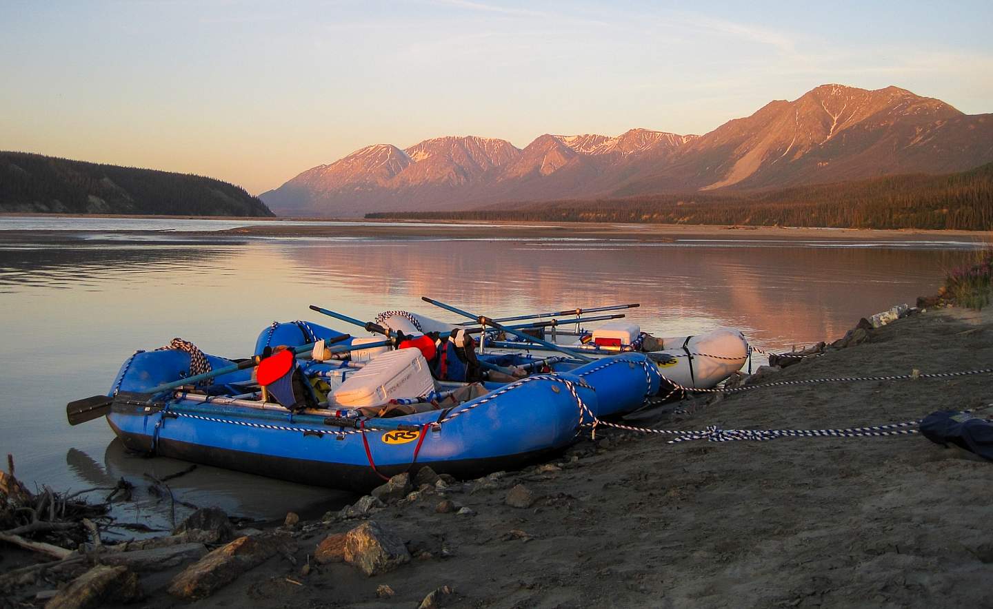 Inflatable rafts filled with camping gear docked on the beach of an Alaskan river.