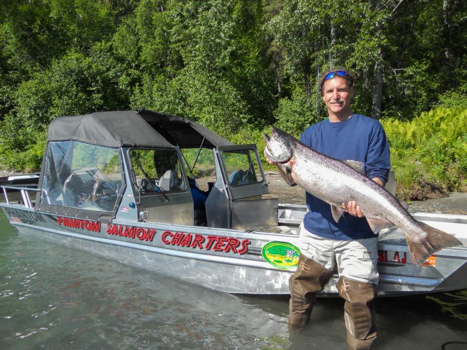 A man in waders stands in front of a fishing boat holding a large fish.