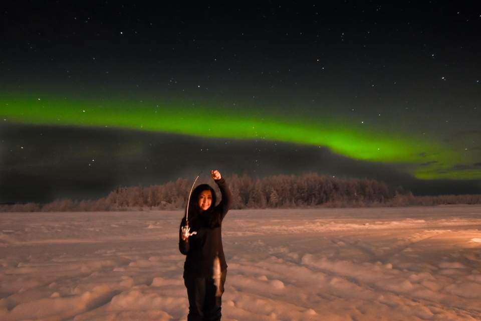 Vibrant green Northern Lights shine above someone who holds up a fish they caught while ice fishing.