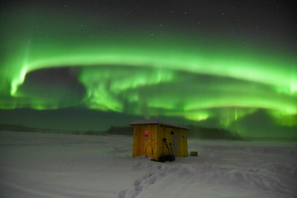 Vibrant green Northern Lights dance above an ice fishing hut.