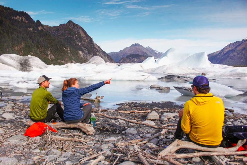 Hikers sit on a rocky beach admiring an ice-choked lagoon.