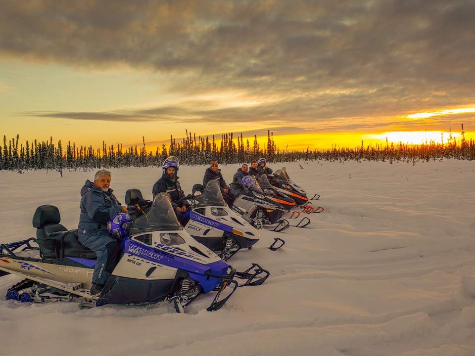 A group of people in a row on their snowmobile with a golden sunset in the background.
