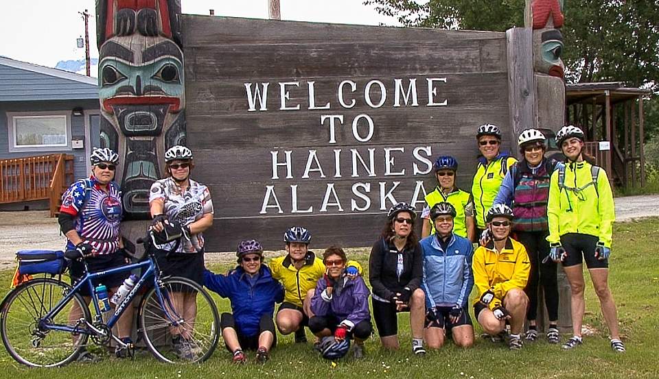 A group of people in cycling gear pose in front of a large wooden sign that says "Welcome to Haines Alaska".
