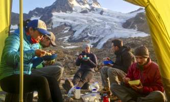 View from inside the cook tent of hungry backpackers enjoying dinner with a glaciated peak behind them Anya Voskresensky
