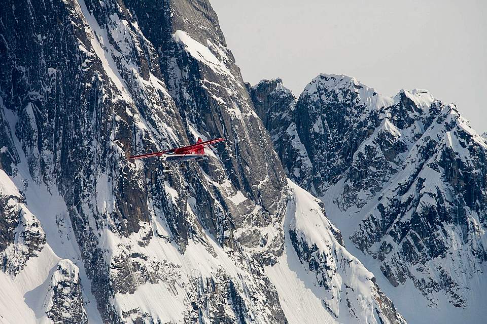 A red bush plane flies close to the face of snow covered Denali.