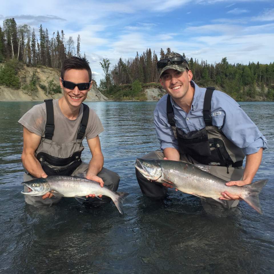 Two men in waders squat in shallow water and hold up fish they caught.