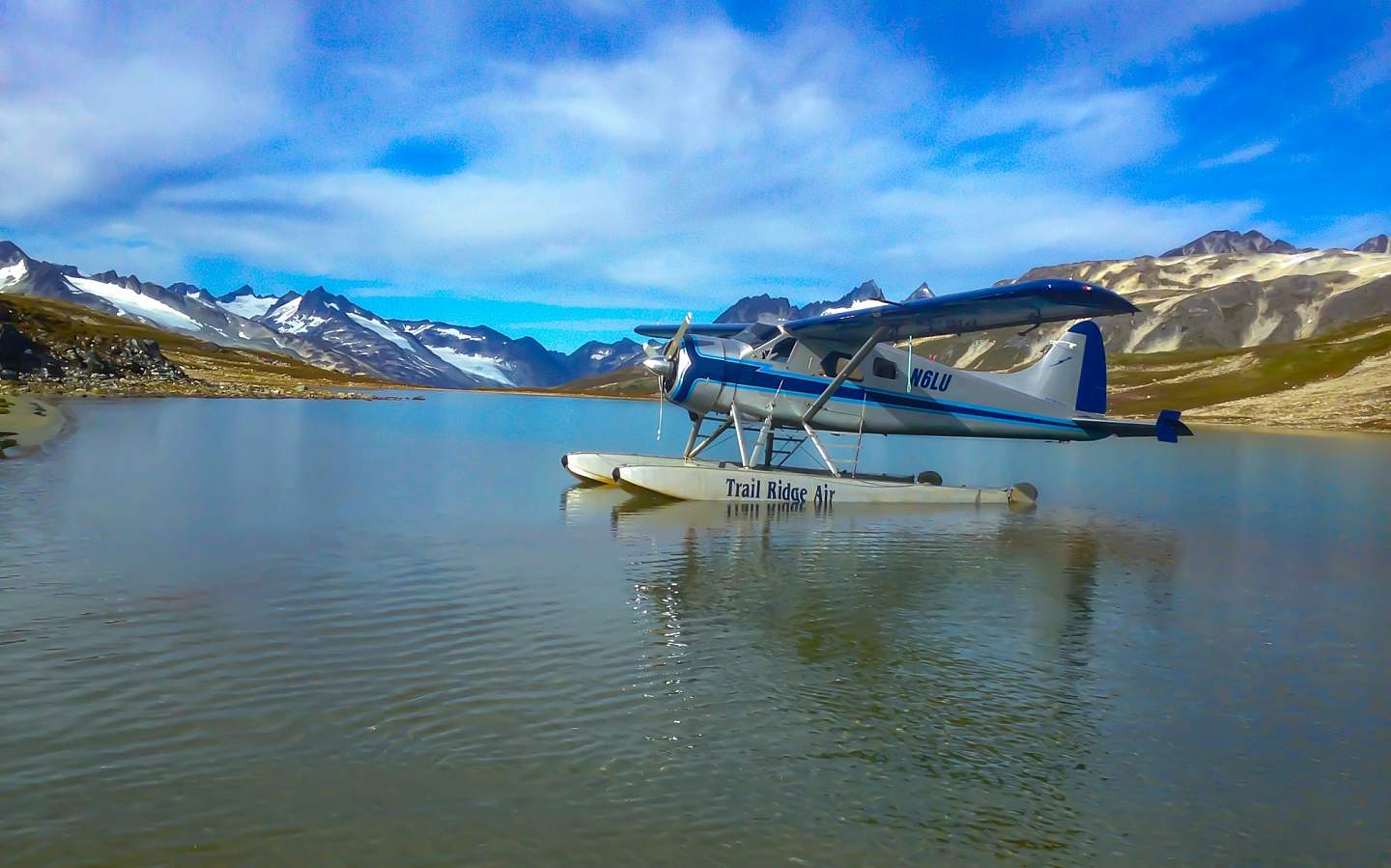 A silver and blue float plane sits on the water with mountains in the background.