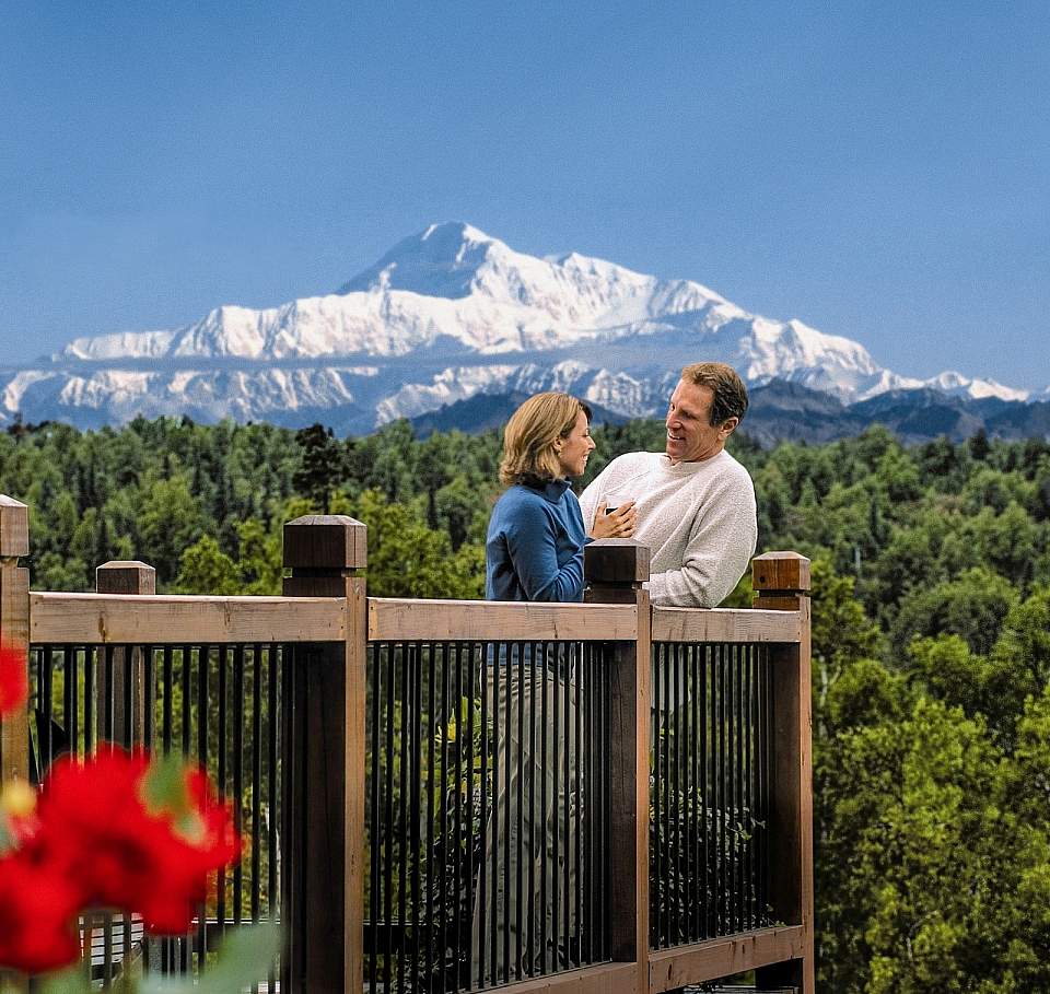 A couple stands on a balcony with Mt. Denali in the background.