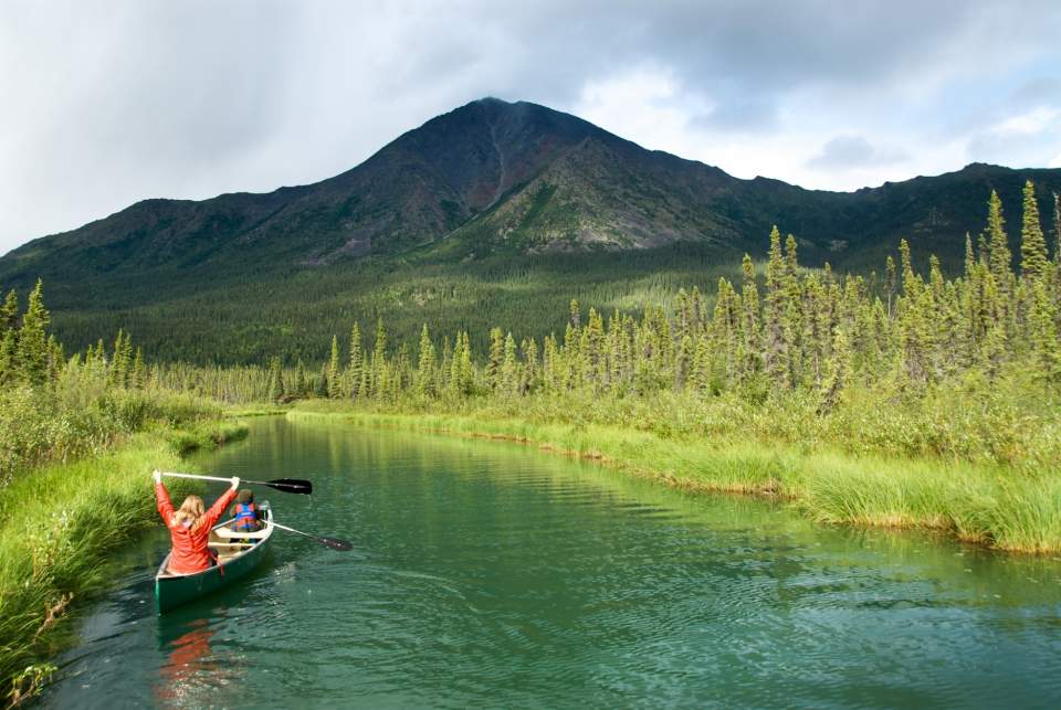 Two people float down a river in a canoe surrounded by lush Alaska landscape.