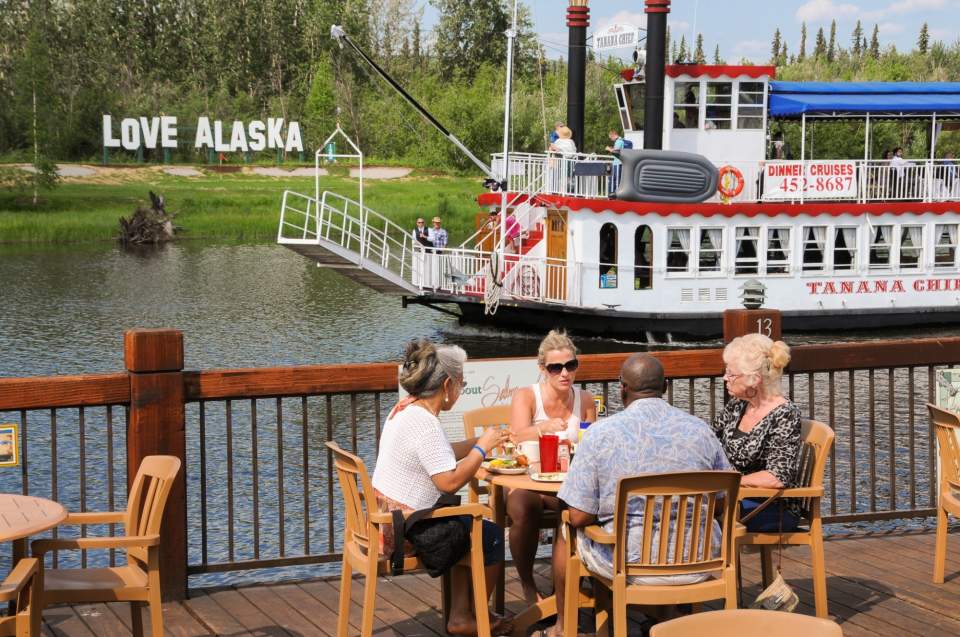 People dining outside on a deck overlooking a river while a river boat sails by.