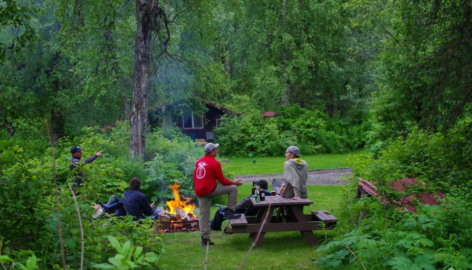 People hanging out around a picnic table and bonfire.