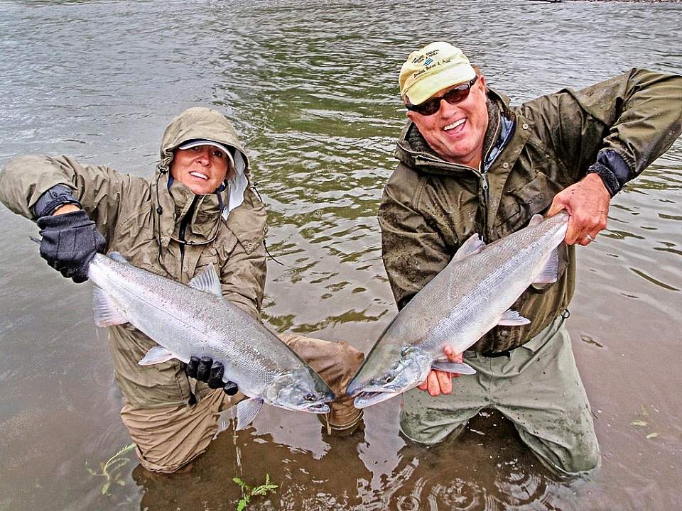 Two people in waders stand in water and hold up fish that they've caught.