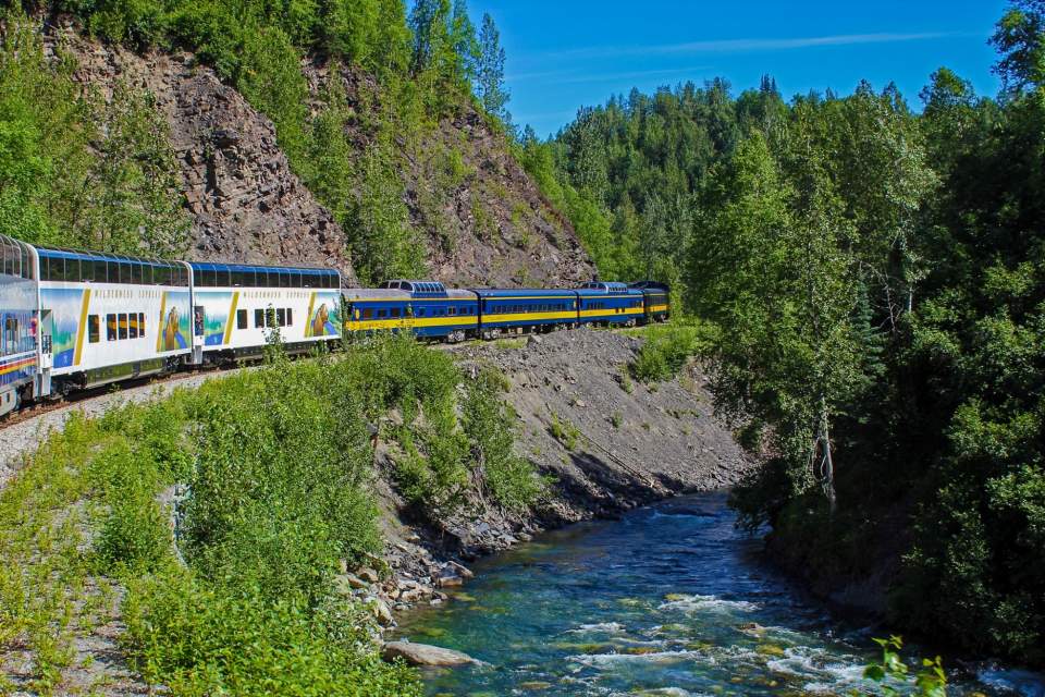 A passenger train travels along a short ridge next to a river surrounded by trees.